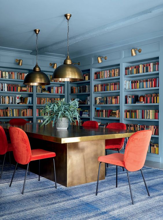 a beautiful stone blue home library and home office with lots of built-in shelves, a large refined desk, red chairs and pendant lamps