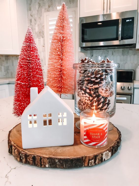 a small Christmas arrangement of a mini house, a jar with snowy pinecones, a candle, pink and red bottle brush Christmas trees