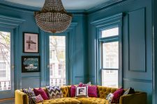 25 a lovely fun and bold stone blue living room with several windows, a mustard corner sofa, colorful and printed pillows, a crystal chandelier