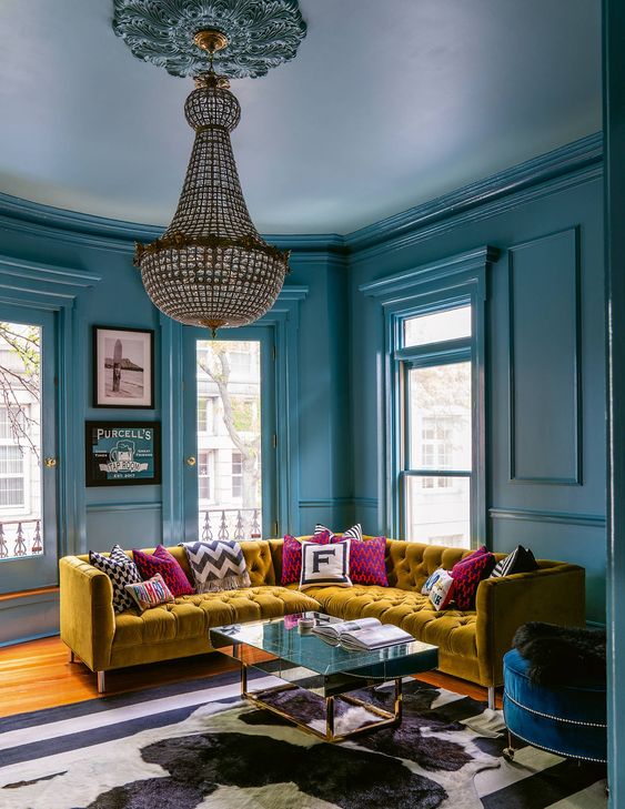 a lovely fun and bold stone blue living room with several windows, a mustard corner sofa, colorful and printed pillows, a crystal chandelier