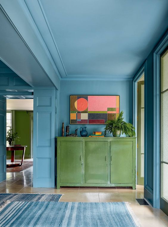 a beautiful and chic stone blue space with much natural light, a green credenza, potted plants and a bold artwork