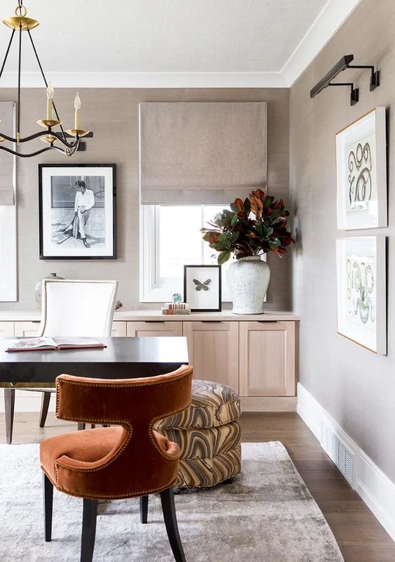 a stylish and neutral modern home office with taupe walls, a light-stained credenza, a black desk and a rust-colored round chair