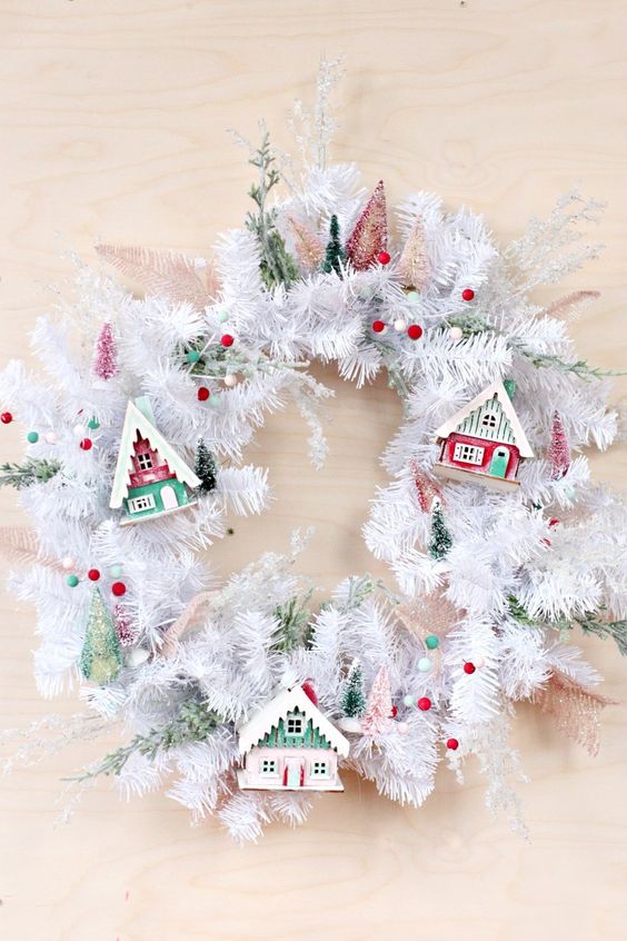 a white evergreen Christmas wreath with colorful pompoms, colorful small houses and bottle brush Christmas trees