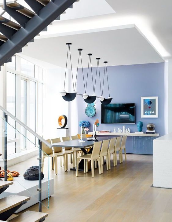 a beautiful white dining space with a very peri accent wall, a large dining table and light-stained chairs, black pendant lamps