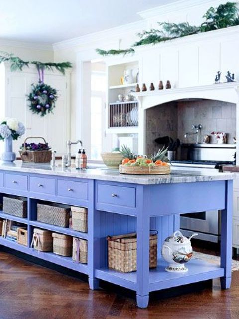 a beautiful white kitchen with an oversized periwinkle kitchen island, baskets for storage, a neutral tile backsplash and lots of evergreens
