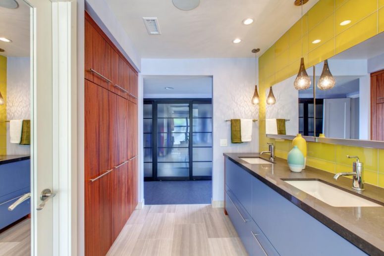 a bold bathroom with a very peri vanity and a yellow tile wall, pendant lamps and a rich-stained storage unit opposite it