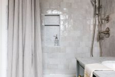 a chic contemporary bathroom clad with light grey and neutral zellige tiles, with a neutral shower curtain and a black vanity