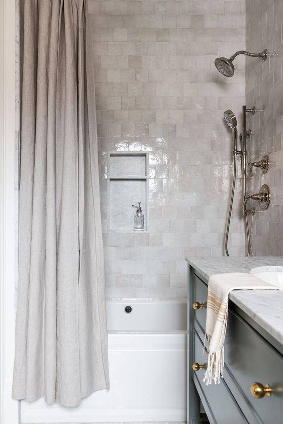 a chic contemporary bathroom clad with light grey and neutral zellige tiles, with a neutral shower curtain and a black vanity