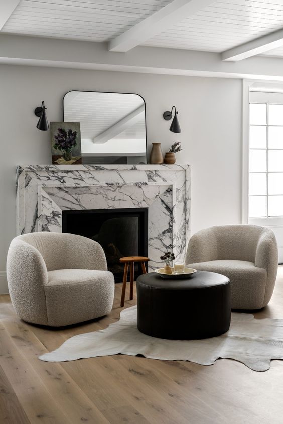 a-chic-contemporary-nook-with-a-white-marble-fireplace-neutral-curved-chairs-a-black-leather-pouf-a-stool-and-some-art-and-decor.jpg