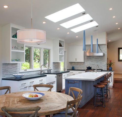 a farmhouse kitchen with skylights, white cabinets and a periwinkle kitchen island, matching pendant lamps over it and a stained wood dining set
