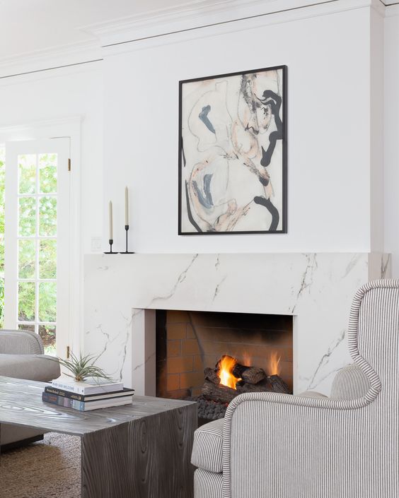a jaw-dropping fireplace nook with a white marble fireplace, striped chairs, a wooden coffee table and a bold artwork