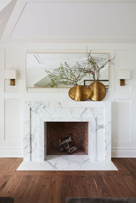 a large non-working fireplace clad with white marble and a mantel, with brass vases with green branches