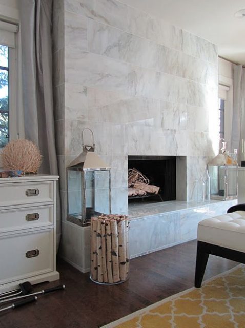 a large non-working fireplace clad with white marble tiles all over, with candle lanterns and firewood in a vase