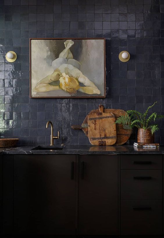 a moody kitchen with black cabinets, a navy zellige tile backsplash, a grey marble countertop and brass and gold fixtures