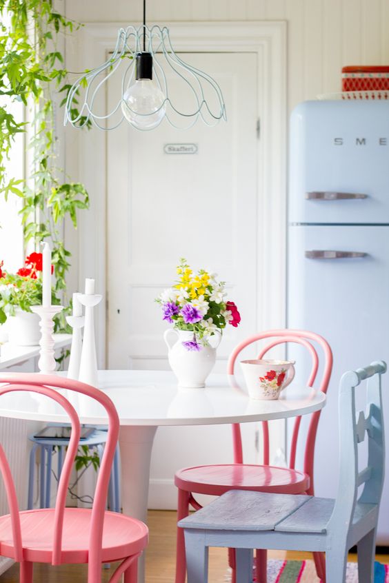 a neutral eat-in kitchen with a periwinkle fridge and chairs, a white table and pink chairs, potted plants and blooms