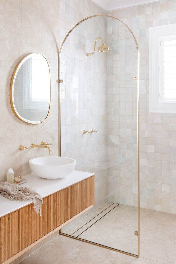 a refined bathroom clad with neutral and pastel zellige tiles in the shower, with a curved space divider and a floating wooden vanity