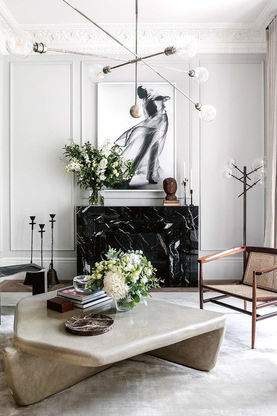 a sophisticated neutral living room with a black marble fireplace, an oversized artwork, a sculptural table and woodne chairs