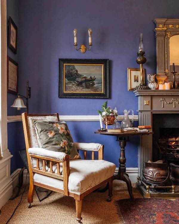 a very peri living room with a grey vintage fireplace, a chic chair and a side table, lots of artworks and lamps is chic and lovely