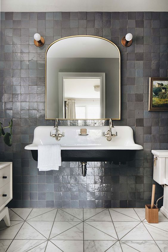 a vintage bathroom with grey zellige tiles, a black floating sink, a curved mirror, brass and gold fixtures and marble tile on the floor