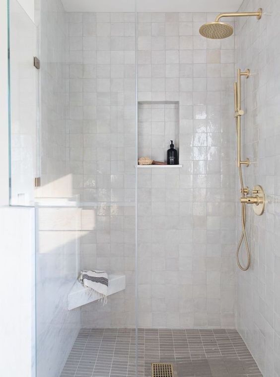 an elegant white shower space completely clad with white zellige tiles, with gold fixtures and a small niche for storage