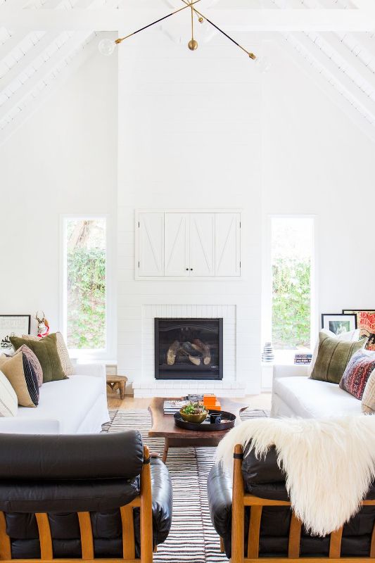 a modern farmhouse living room in white, with white sofas and black leather chairs, a fireplace and a TV behind white barn doors