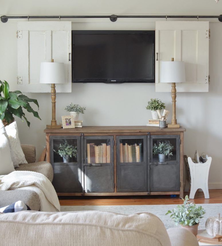 a neutral farmhouse living room with neutral sitting furniture, a TV on the wall hidden behind white barn doors and a metal and wood storage unit