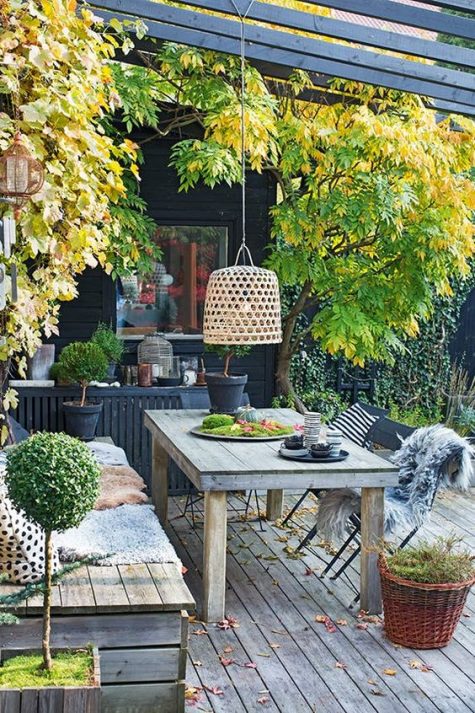 a relaxed Scandinavian deck with a built in bench and a table, a pendant lamp, potted greenery and some trees
