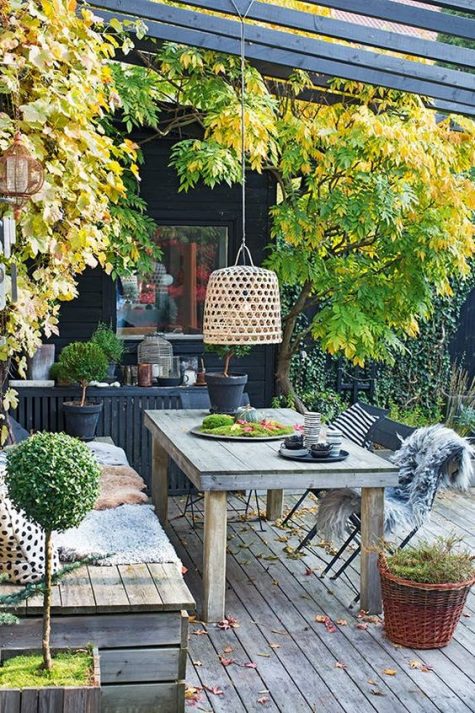 a relaxed Scandinavian deck with a built-in bench and a table, a pendant lamp, potted greenery and some trees