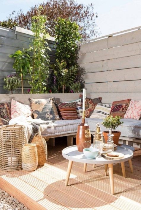 an inviting deck with a corner pallet bench and printed pillows, round tables, candle lanterns and potted greenery and climbing plants