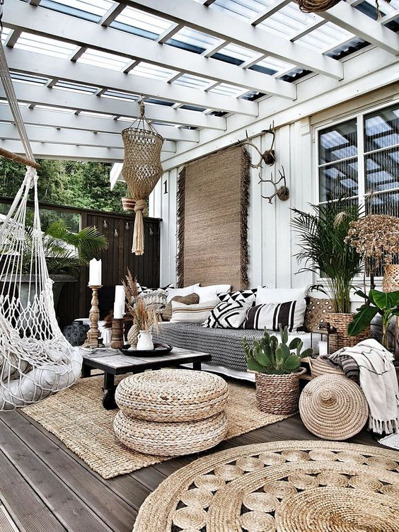 a boho outdoor living room with a glass roof, a large sofa, jute rugs and poufs, woven chairs and printed pillows