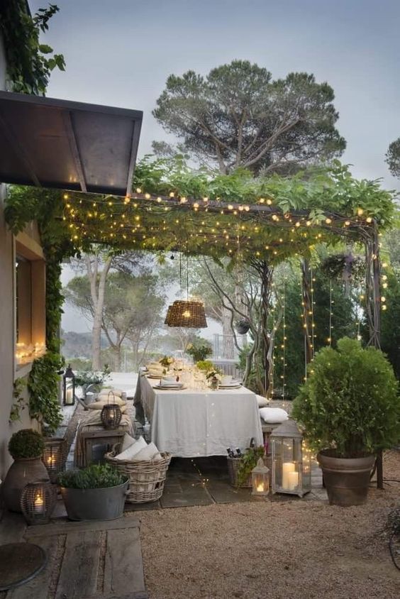 a lovely Provence outdoor dining space with elegant dining furniture, white linens, woven pendant lamps, greenery and lots of lights