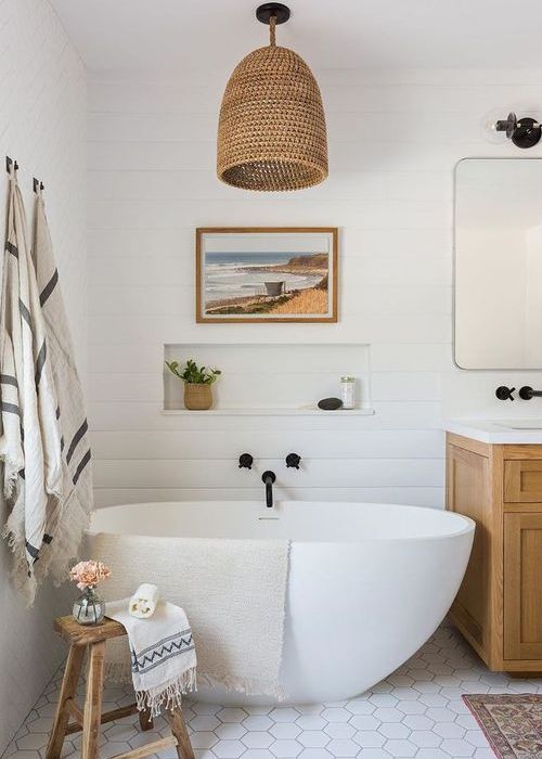 a white bathroom clad with shiplap, with hex tiles on the floor, an oval tub, a stained vanity, a woven pendant lamp and printed textiles