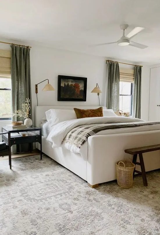 a chic and welcoming bedroom with a creamy upholstered bed, neutral bedding, chic nightstands, a wooden bench, baskets and green curtains