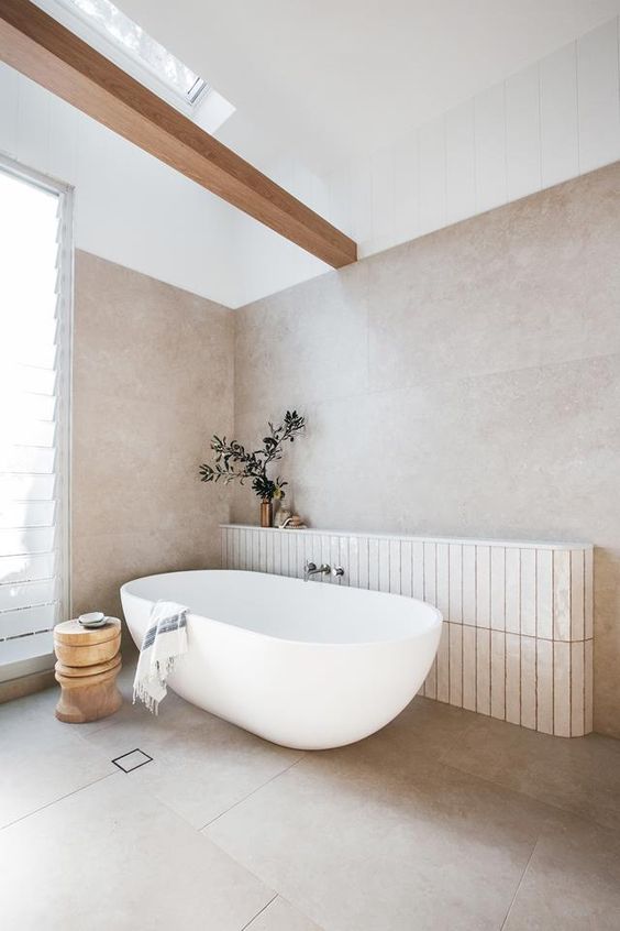 a beautiful bathroom with blush limewashed walls, an oval tub and a wooden side table is light-filled through the window and the skylight