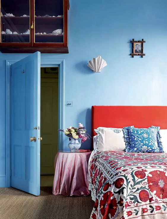 a beautiful blue bedroom with a coquelicot bed and bright printed bedding with the same shades, a vintage nightstand with blooms