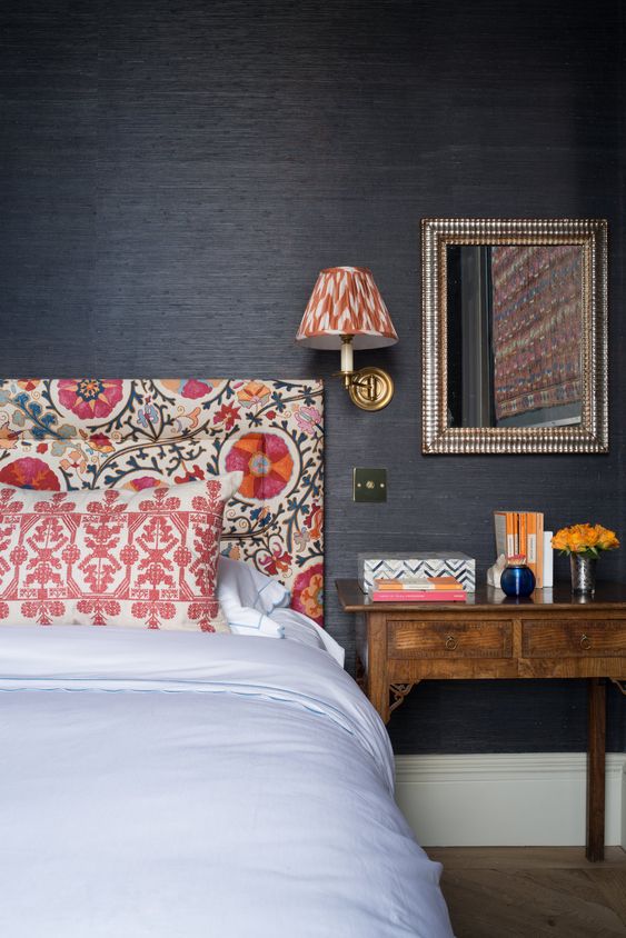 a bedroom with black grasscloth wallpaper walls, a bold printed bed and a wall sconce, a stained nightstand
