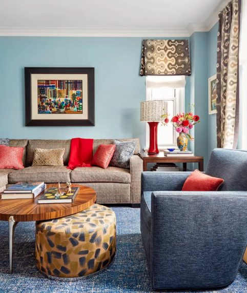 a blue living room with grey and tan seating furniture, printed curtains and an ottoman, bold coquelicot pillows and a lamp for a touch of color