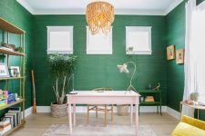 a bright home office with green grasscloth wallpaper, a pink desk, a yellow chair, a beaded chandelier and a brass storage unit