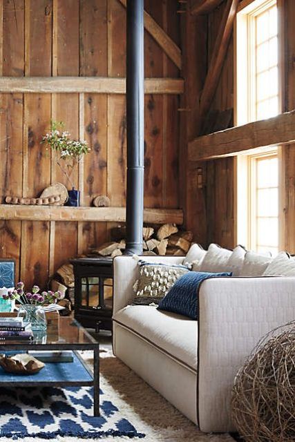 a cabin with rough wooden walls and beams that bring coziness to the space, a black metal hearth and a white sofa with pillows