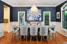 a catchy dining room with navy grasscloth wallpaper, white wainscoting, a grey dining table, grey chevron chairs, a bold artwork and a chic chandelier