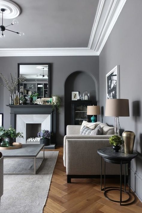 a chic living room with slate grey walls, neutral seating furniture and grey and black tables, a faux fireplace and lamps