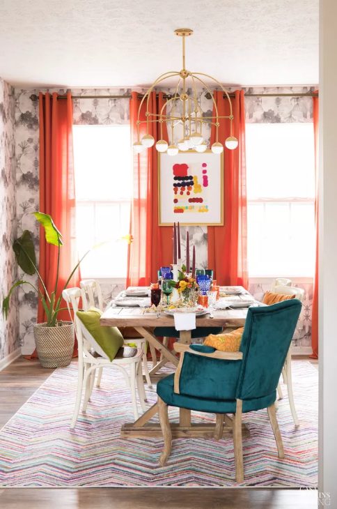a colorful dining space with printed wallpaper, a wooden table, mismatching chairs, coquelicot curtains and matching touches on the table