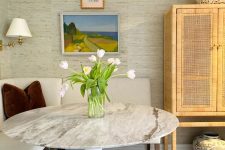 a cozy dining nook with neutral grasscloth wallpaper, a creamy banquette seating, a round marble table and a cane storage unit