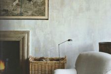 a cozy space with grey limewashed walls, a fireplace, a creamy chair, a basket with firewood and an artwork