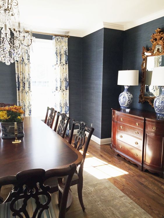 a moody and refined vintage dining room with black grasscloth wallpaper, dark stained vintage furniture, a mirror in an ornated frame and a crystal chandelier