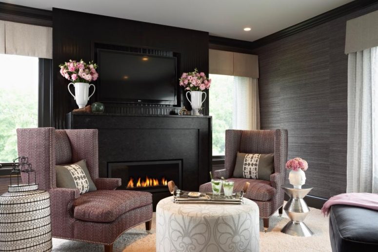 a moody living room with graphite grey grasscloth wallpaper, a built-in fireplace with a black surround, mauve chairs and neutral ottomans