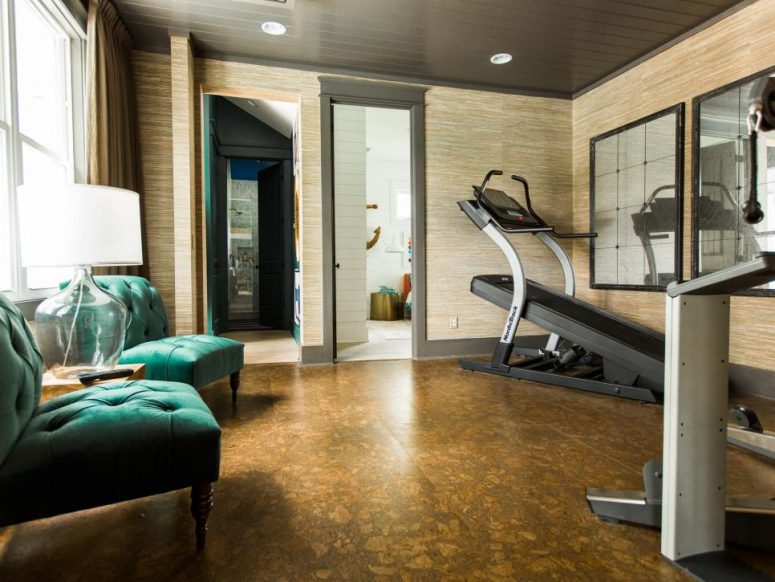 a small and cozy home gym styled with neutral grasscloth wallpaper, a stone floor, green tufted chairs and a table lamp
