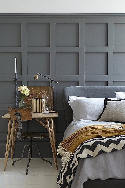 a stylish bedroom with slate grey paneled walls, a slate grey upholstered bed, neutral and printed bed, a wooden nightstand and a vintage chair