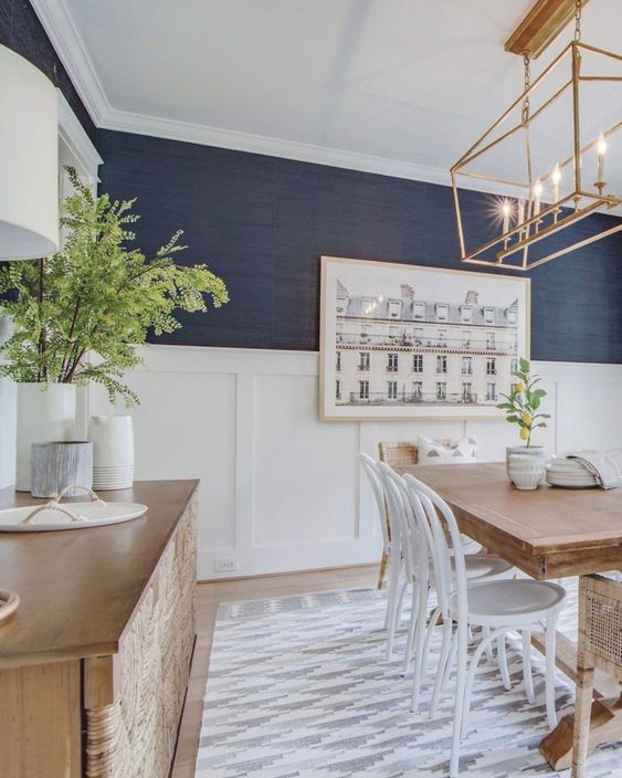 a stylish dining room with navy grasscloth wallpaper, white wainscoting, a stained wooden table and a credenza and white chairs