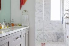 a stylish neutral bathroom clad with white marble tiles, with a grey vanity, a bold striped coquelicot rug and a matching hanging with long fringe